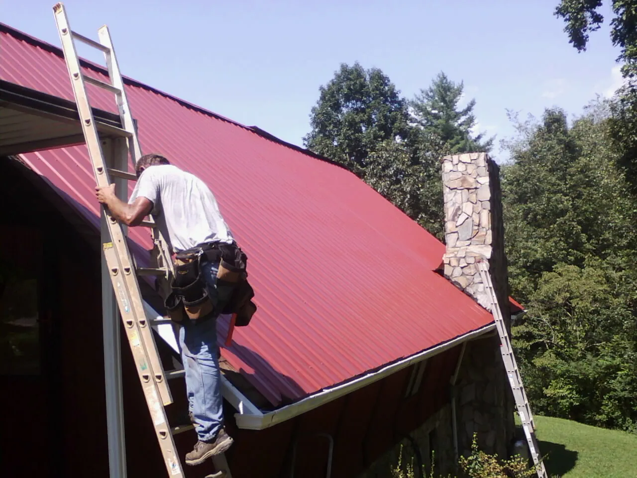 Roofer on a ladder beside a red metal roof and stone chimney — Soffit & Fascia Repair by Roofing Brothers in Emmitsburg